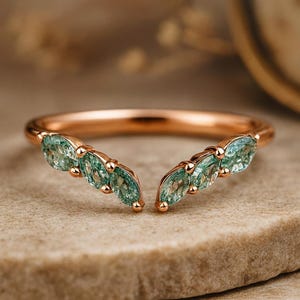 Könnte beinhalten: Roségoldener Ring mit geschwungenem Design. Der Ring hat fünf marquiseförmige, hellgrüne Edelsteine auf jeder Seite. Die Edelsteine sind in einem zarten, offenen Design gefasst. Der Ring wird auf einer neutralen Oberfläche präsentiert.