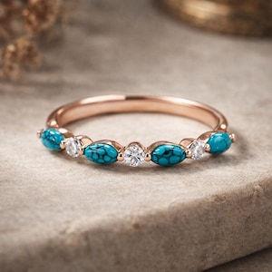 May include: Rose gold ring with alternating turquoise and clear gemstones. The turquoise stones are oval, and the clear stones are round. The ring is displayed on a neutral surface, highlighting the jewellery's design and colour.