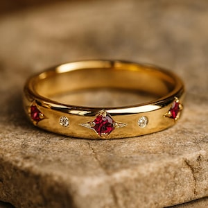 May include: A gold ring with three red square gemstones and two small round diamonds. The ring has a smooth, polished surface and is set against a neutral background. The ring is a piece of jewelry.