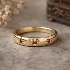 Peut inclure: Une bague dorée ornée de trois motifs d'éclats d'étoiles, chacun serti d'une petite pierre précieuse rouge ronde. La bague a une surface lisse et polie et un design de bande classique. La bague est un bijou.