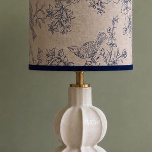 Op de afbeelding: Een decoratieve tafellamp met een crème keramische voet en een stoffen lampenkap. De lampenkap heeft een blauw en wit vogel- en bloemenontwerp, met een donkerblauwe rand. De lamp heeft een messingkleurige hals.