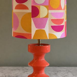Puede incluir: Una lámpara de mesa con una base de cerámica naranja brillante que presenta una silueta escalonada en forma de reloj de arena. La pantalla cilíndrica de tela muestra un patrón geométrico repetitivo de círculos y semicírculos en tonos rosa, naranja, amarillo y blanco.