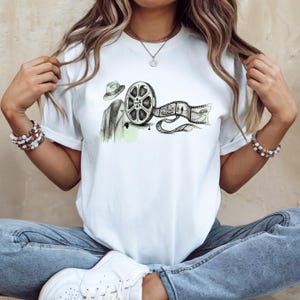May include: White t-shirt featuring a film reel graphic with a fedora and coat. The graphic is in black and white with a touch of green. The film strip shows a landscape with two figures. The shirt is a classic crew neck.