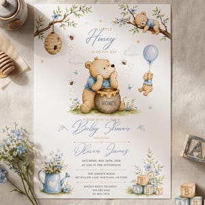 Peut inclure: Une carte d'invitation pour une fête prénatale avec une illustration à l'aquarelle d'un ours en chemise bleue avec un pot de miel, des abeilles et des fleurs bleues. Le texte indique A Little Honey Is On The Way, Baby Shower, Oliver James.