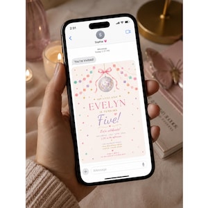 May include: A smartphone displaying a digital birthday invitation on its screen. The invitation features a light pink background with a disco ball, colorful bunting, and text that reads: Our little star Evelyn is turning Five! Let's celebrate.