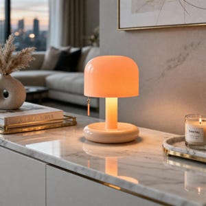 Peut inclure: Une lampe de table pêche avec un abat-jour arrondi et une base cylindrique. La lampe est allumée, émettant une lueur chaude. La lampe est sur une surface en marbre, avec une bougie et des objets décoratifs en arrière-plan.
