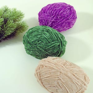 May include: Three balls of soft, velvety yarn in shades of purple, green, and beige. The yarn is arranged diagonally, with a sprig of green foliage in the upper left corner. The yarn appears to be a plush, textured material, suitable for knitting or crocheting projects.