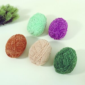 May include: A collection of six skeins of yarn in various colors: brown, light green, purple, beige, and dark green. The yarn is arranged on a light green surface. The yarn appears to be soft and textured.