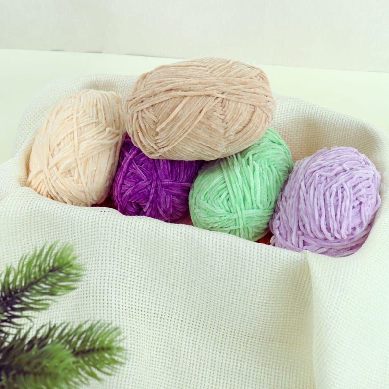 May include: A collection of colorful yarn balls in a white basket. The yarn colors include beige, purple, light green, and lavender. The yarn is soft and fluffy, perfect for knitting or crocheting projects. The basket is made of a woven fabric.