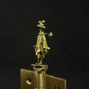 May include: A brass-colored hinge with a decorative frog figure on top. The frog is wearing a scarf and cape. The hinge has a square shape with screw holes, set against a dark background.