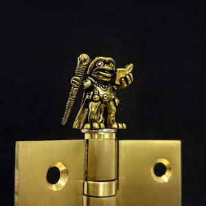 Pode incluir: Uma estatueta de rã cor de latão, segurando um bastão e um livro, em cima de uma dobradiça de latão. A rã é detalhada com uma capa e cinto. O fundo é preto.