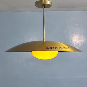 May include: A brass-colored pendant light fixture with a circular canopy and a long, slender rod. The light has a wide, shallow, saucer-shaped shade with a yellow, semi-circular light source. The text "UKBRASSLIGHTING" is printed on the shade.