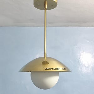 May include: A brass pendant light fixture with a white globe shade. The fixture has a round canopy and a long, slender rod. The shade is a half-dome shape with the text "UKBRASSLIGHTING" on the edge.