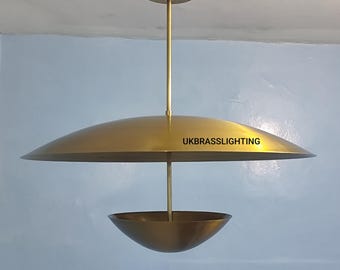 May include: A gold-colored, modern pendant light fixture. The fixture has a large, shallow, disc-shaped shade and a smaller, bowl-shaped element suspended below. The text "UKBRASSLIGHTING" is printed on the shade.
