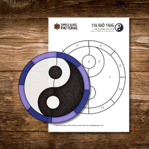 Può includere: Un modello di vetrata Yin e Yang con un design in bianco e nero, circondato da un bordo blu e viola. Il modello è accanto a un foglio con un diagramma e il testo "Yin and Yang". Dimensioni: 19 x 19 cm.