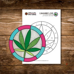May include: A stained glass pattern featuring a cannabis leaf design. The pattern is circular, with a green leaf at the center, surrounded by a teal and pink border. The design is on a white sheet with the text "CANNABIS LEAF".