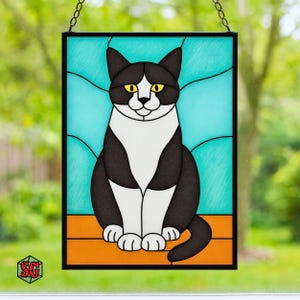 K&ouml;nnte beinhalten: Buntglasfenster mit einer schwarz-wei&szlig;en Katze mit gelben Augen, vor einem hellblauen Hintergrund. Die Katze sitzt auf einer orangefarbenen Oberfl&auml;che, ist schwarz umrandet und an einer Kette aufgeh&auml;ngt. Ein dekoratives St&uuml;ck f&uuml;r Katzenliebhaber.