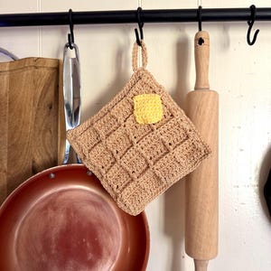 May include: A tan crochet potholder shaped like a waffle, with a yellow square of butter on top. The potholder is hanging from a black metal bar with other kitchen utensils, including a copper pan and a wooden rolling pin.