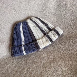 Op de afbeelding: Een handgemaakte gehaakte beanie met strepen in marineblauw, wit en grijs. De muts heeft een omgeslagen rand en is gemaakt van zacht, getextureerd garen. Deze wintermuts is ideaal voor koud weer en heeft een diameter van ongeveer 20 cm.