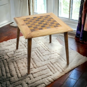 Puede incluir: Pequeña mesa de ajedrez cuadrada de madera con tablero de ajedrez. La mesa tiene cuatro patas delgadas y cónicas y está colocada sobre una alfombra color crema. El tablero de ajedrez está hecho de cuadrados de madera clara y oscura.