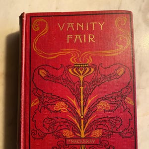 May include: A hardcover book titled "Vanity Fair" with a red cover and gold lettering. The cover features an ornate gold and black floral design. The author's name, Thackeray, is printed at the bottom.