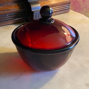 May include: A small, round, covered dish with a deep red glass lid and a black base. The lid has a black knob on top. The dish is sitting on a white marble surface.