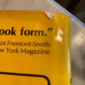 以下が含まれることがあります： 「ook form.」のテキストと「Fremont-Smith」と「New York Magazine」の名前が書かれた、本の黄色の表紙のクローズアップ。 本の右上隅が少し破損しています。