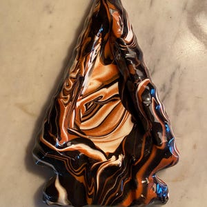May include: A ceramic arrowhead with a marbled design in black, orange, and white. The arrowhead is triangular with a notched base and a glossy finish. The colors swirl together, creating a unique pattern.