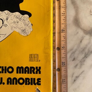 以下が含まれることがあります： 黄色い背景に、帽子をかぶった人物の白黒カリカチュアが描かれたヴィンテージポスター。ポスターには「CHO MARX J. ANOBILE」の文字とアーティスト名「HIRSCHFELD」が記載されています。木製の定規がポスターの横に置かれています。