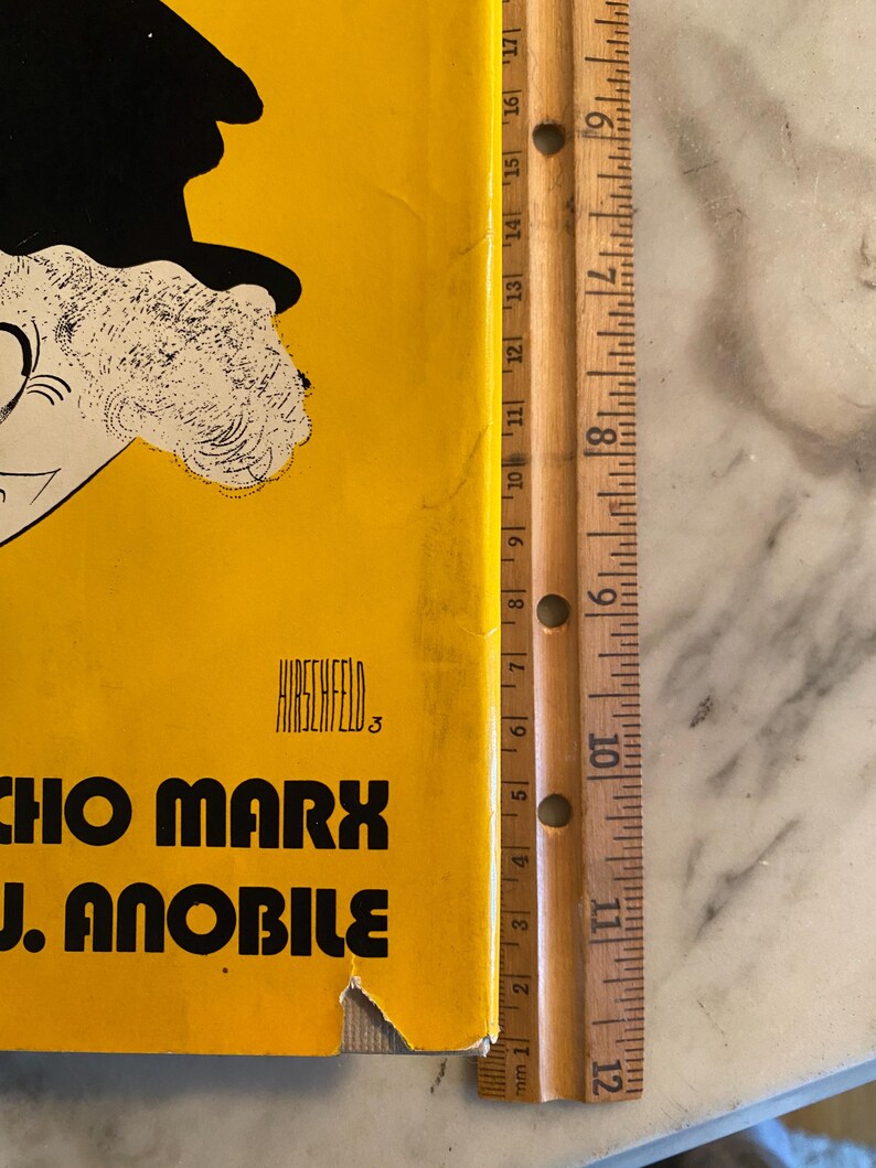 以下が含まれることがあります： 黄色い背景に、帽子をかぶった人物の白黒カリカチュアが描かれたヴィンテージポスター。ポスターには「CHO MARX J. ANOBILE」の文字とアーティスト名「HIRSCHFELD」が記載されています。木製の定規がポスターの横に置かれています。
