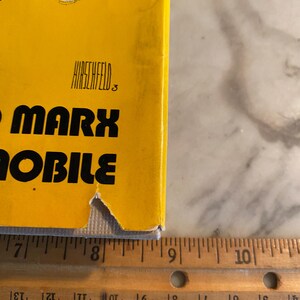 以下が含まれることがあります： 大きな黒い文字で「MARX MOBILE」というタイトルが書かれた黄色の本の表紙。著者の名前「HIRSCHFELD」がタイトルの上にあります。木の定規が本の隣に置かれ、センチメートル単位の測定値が表示されています。