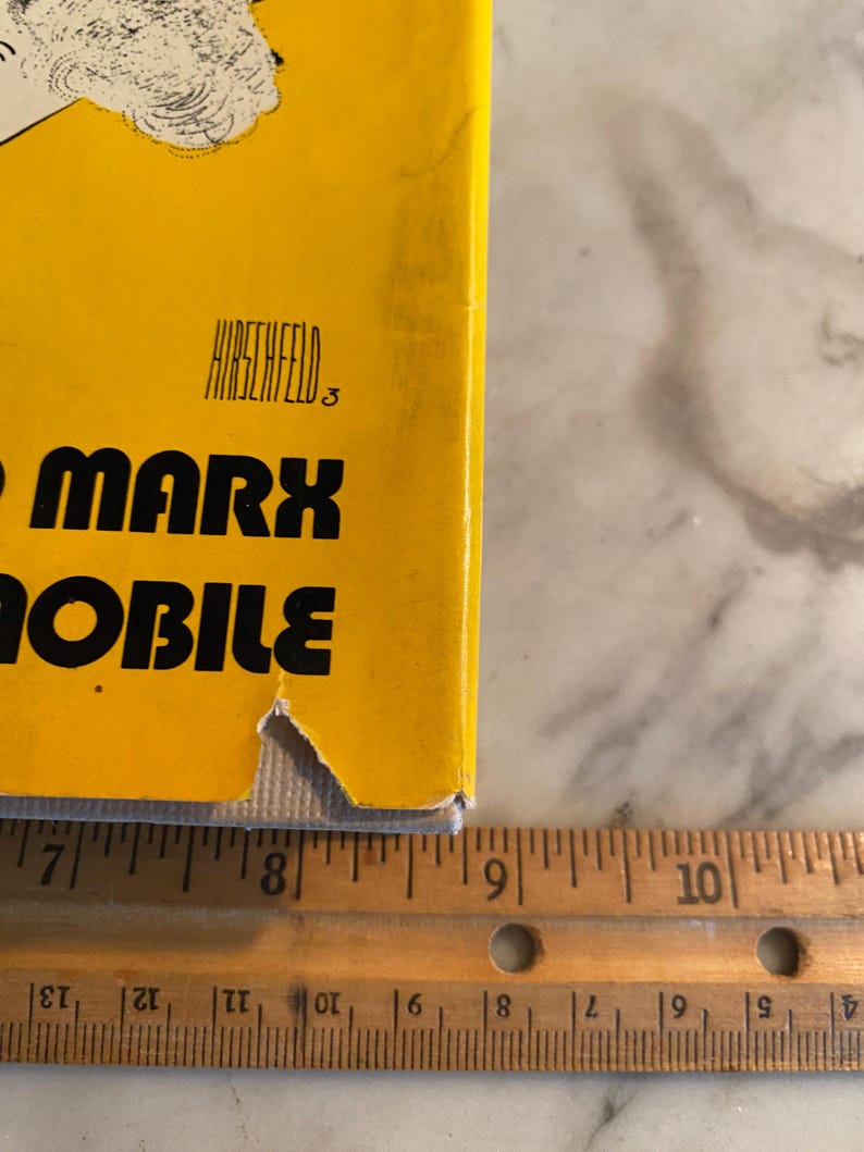 以下が含まれることがあります： 大きな黒い文字で「MARX MOBILE」というタイトルが書かれた黄色の本の表紙。著者の名前「HIRSCHFELD」がタイトルの上にあります。木の定規が本の隣に置かれ、センチメートル単位の測定値が表示されています。