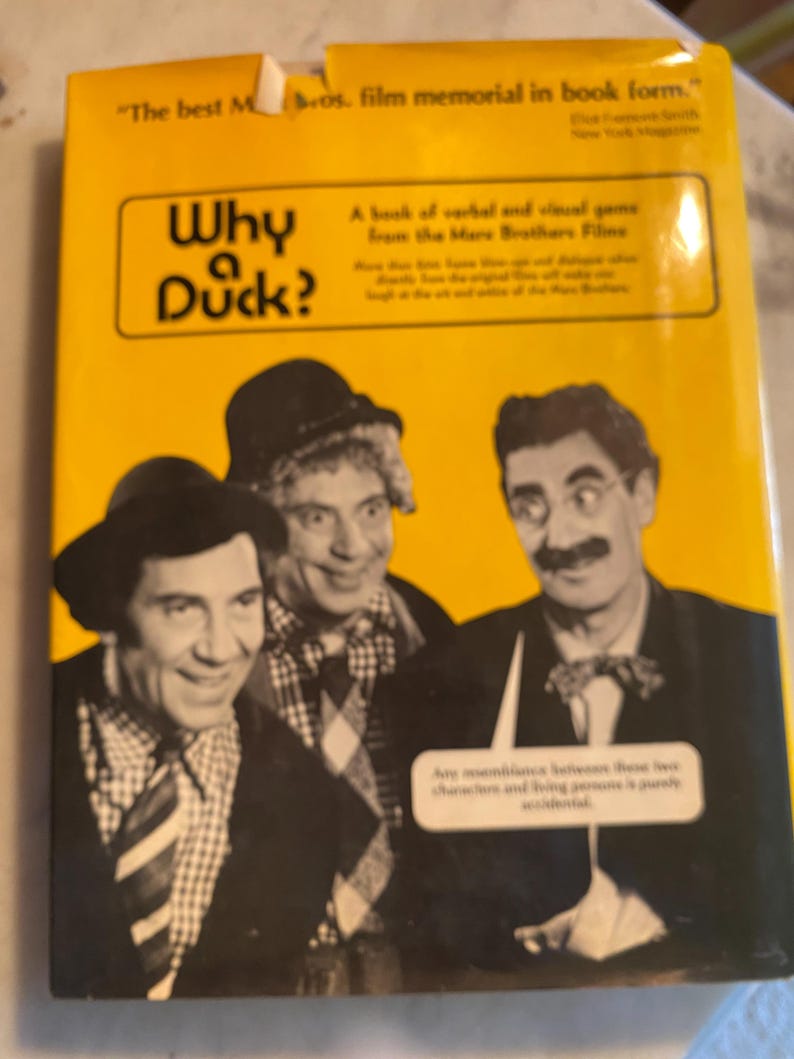 以下が含まれることがあります： 「Why Duck?」のタイトルとマルクス兄弟の写真が描かれた黄色の本の表紙。表紙には、映画の記念碑であり、マルクス兄弟の映画からの言葉とビジュアルゲームであることについてのテキストが含まれています。