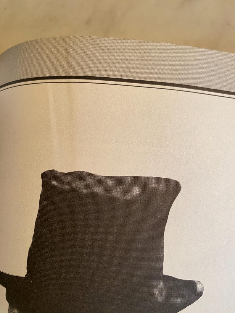 以下が含まれることがあります： 黒いシルクハットの白黒イラスト。帽子は高く円筒形のクラウンと幅広の平らなつばが特徴です。画像はクローズアップで、帽子の形と質感を強調しています。