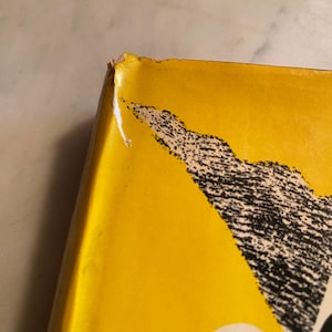 以下が含まれることがあります： 鮮やかな黄色の表紙と白黒のグラフィックデザインが特徴的な本の角のクローズアップ。角には摩耗があり、端に小さな破れがあります。