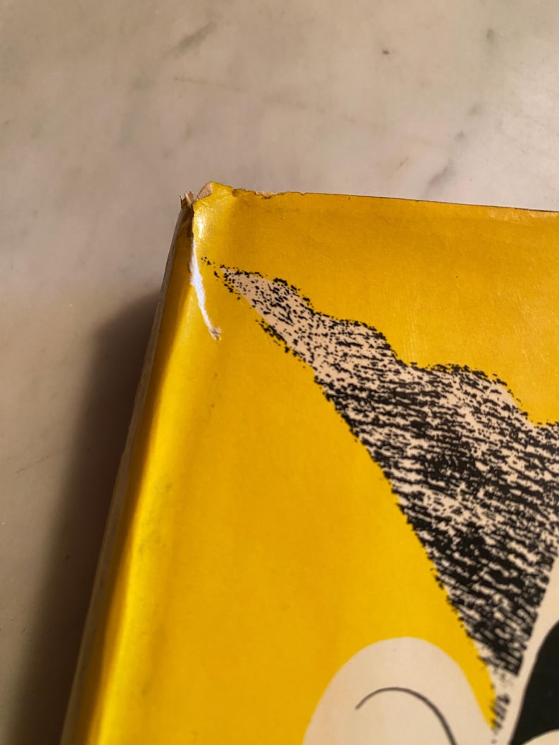 以下が含まれることがあります： 鮮やかな黄色の表紙と白黒のグラフィックデザインが特徴的な本の角のクローズアップ。角には摩耗があり、端に小さな破れがあります。