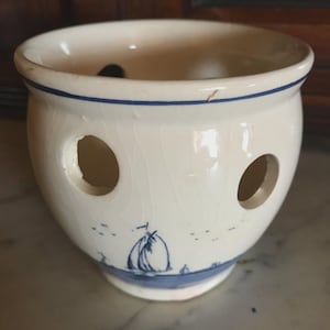 May include: Cream-colored ceramic onion keeper with three large holes and a blue sailboat design. The rim and base feature a blue stripe. The keeper has a round shape and is sitting on a marble surface.