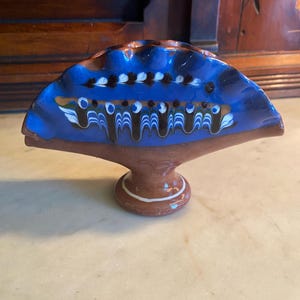 May include: A ceramic fan-shaped decorative piece with a blue top and brown base. The blue section features white, black, and brown painted designs. The base is brown with a white accent ring. This decorative item is likely for display.