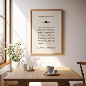May include: A framed print with the text "Tomorrow is a New Day" and a quote by Ralph Waldo Emerson. The print is in a wooden frame and is displayed on a table with a vase of white flowers and a tea set.