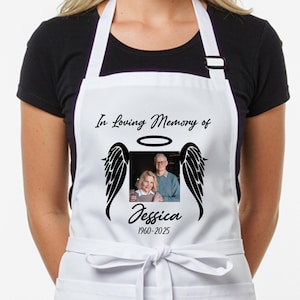 May include: White apron with the text "In Loving Memory of" above a photo of two people, with angel wings and a halo. The name "Jessica" and the years "1960-2025" are below the photo. The apron is tied at the waist.