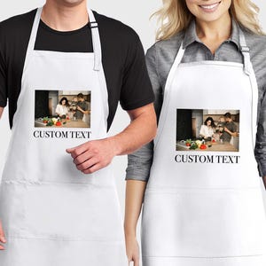 May include: White aprons with a black neck strap. Each apron has a rectangular image of a family in a kitchen and the words "CUSTOM TEXT" printed below the image. The aprons are worn by two people.