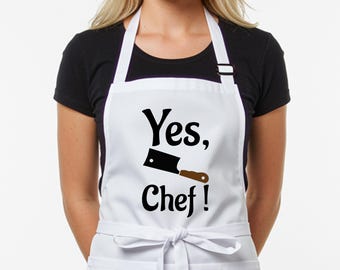 May include: White apron with the text "Yes, Chef!" in black script. A black cleaver graphic with a brown handle is centered below the text. The apron has a neck strap and waist ties.