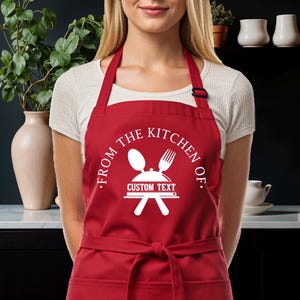 May include: A bright red apron featuring a white graphic print of a spoon, a fork, and a covered serving dish. The text reads FROM THE KITCHEN OF and CUSTOM TEXT. The apron has a neck strap with a black adjustable buckle and a waist tie.
