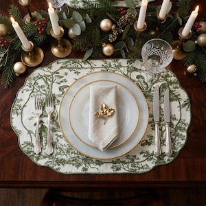 Può includere: Una tovaglietta smerlata con motivo toile verde e bianco, due piatti bianchi con bordi dorati, un tovagliolo di lino beige, un portatovagliolo a foglia dorata, due forchette, un coltello d'argento e un calice di vetro su un tavolo.
