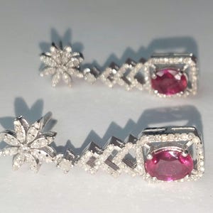 Könnte beinhalten: Ein Paar silberne Ohrringe mit einem floralen Stecker, einem geometrischen Design und einer quadratischen Fassung mit einem leuchtend roten Edelstein. Die Ohrringe sind mit kleinen, funkelnden Akzentsteinen besetzt.