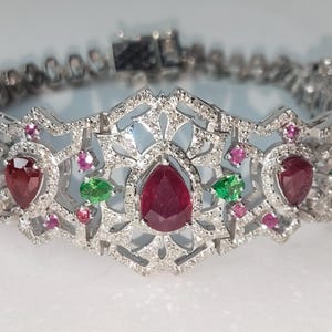 Könnte beinhalten: Ein silbernes Armband mit dekorativem Design, das birnenförmige rote Edelsteine, kleine grüne Edelsteine und winzige rosa Edelsteine aufweist. Das Armband ist mit kleinen, klaren Steinen besetzt.
