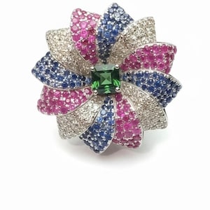 Könnte beinhalten: Ein blütenförmiger Ring mit einem quadratischen grünen Edelstein in der Mitte. Die Blütenblätter bestehen aus abwechselnden Abschnitten aus rosa, blauen und klaren Edelsteinen, wodurch ein lebendiges Design entsteht. Der Ring ist auf einem silbernen Band gefasst.