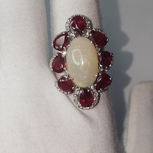 Könnte beinhalten: Ein silberfarbener Ring mit floralem Design. Der Ring hat einen großen, ovalen, cremefarbenen Opal in der Mitte, umgeben von kleineren, tropfenförmigen, tiefroten Edelsteinen. Der Ring ist mit kleinen, klaren Akzentsteinen besetzt und wird auf weißem Untergrund präsentiert.