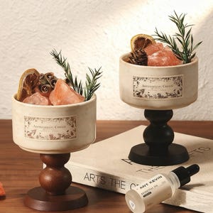 Puede incluir: Dos candelabros de aromaterapia con un cuenco color crema, llenos de cristales rosas, cítricos secos y vegetación. Los candelabros tienen una base de madera y una etiqueta que dice "Aromatherapy Candle". También hay una botella de aceite esencial.