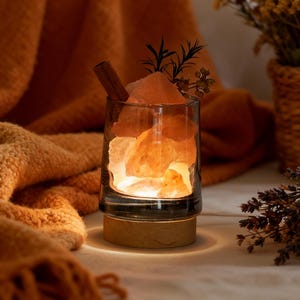May include: A Himalayan salt lamp in a clear glass with a wooden base, filled with pink salt crystals. A cinnamon stick and sprigs of greenery sit atop the lamp. An orange blanket and a basket of dried flowers are in the background.